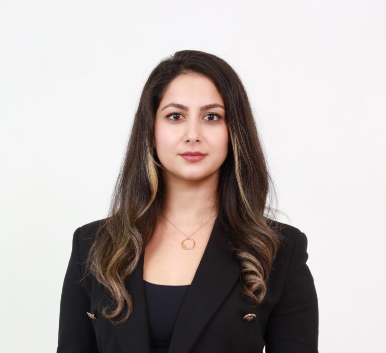 A professional headshot of Ghazal Hamedani, a woman with long dark hair and highlights, wearing a black blazer and a gold necklace, set against a clean background. To the left, a decorative image of dried plant stems is overlaid with the text "NEW BLOG POST" in a circular design. Below, the title reads "Why It’s Time to Revisit Your Unanimous Shareholder Agreement (USA)" followed by "by Ghazal Hamedani" and the website "www.kalfalaw.com" in smaller font. The design features soft beige and white abstract shapes.
