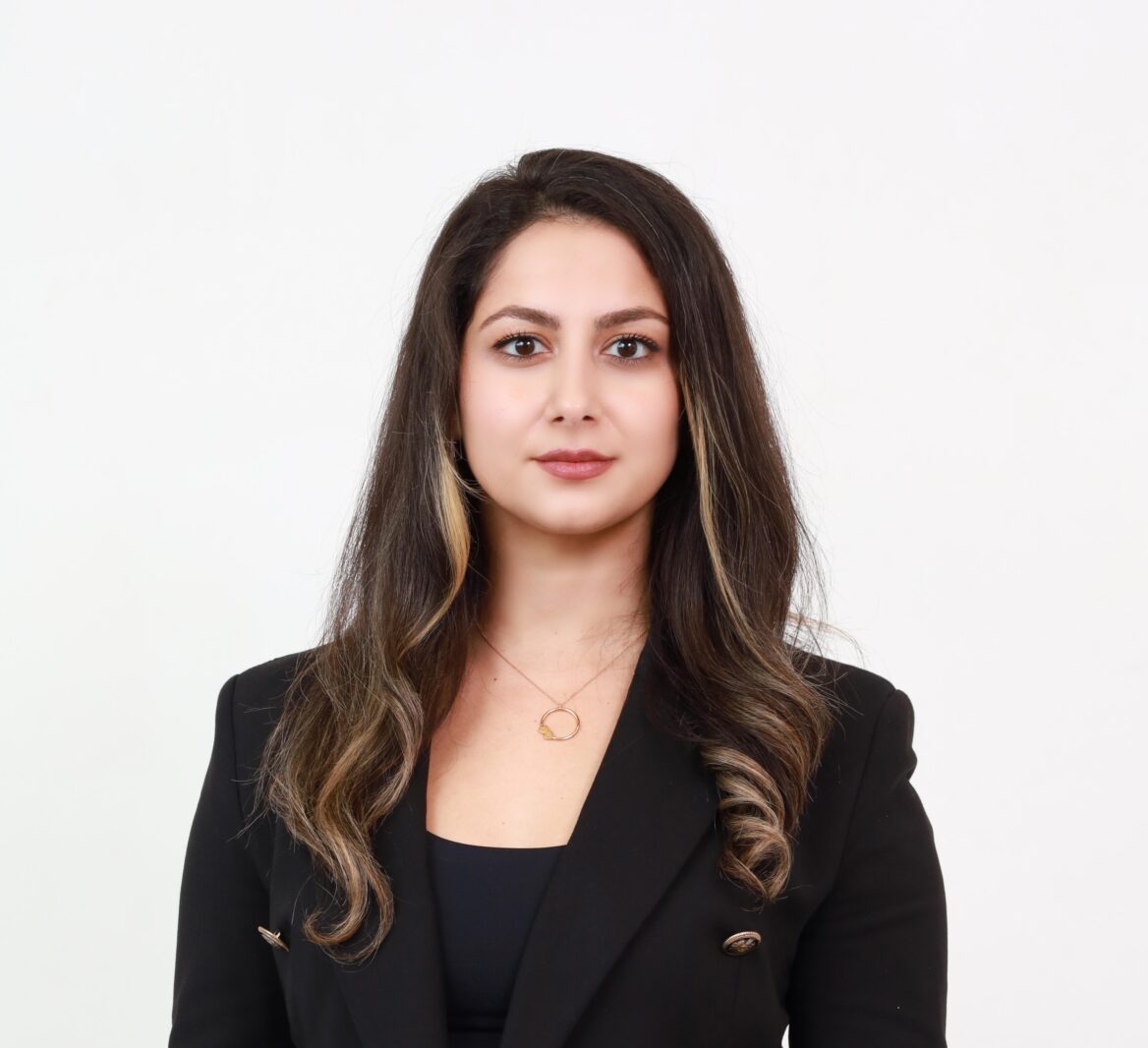 A professional headshot of Ghazal Hamedani, a woman with long dark hair and highlights, wearing a black blazer and a gold necklace, set against a clean background. To the left, a decorative image of dried plant stems is overlaid with the text "NEW BLOG POST" in a circular design. Below, the title reads "Why It’s Time to Revisit Your Unanimous Shareholder Agreement (USA)" followed by "by Ghazal Hamedani" and the website "www.kalfalaw.com" in smaller font. The design features soft beige and white abstract shapes.